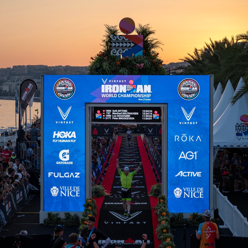 A man in a neon triathlon kit raises his arms up as he crosses the blue finishing arch of the IMWC Nice 2023 race at dusk.